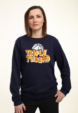 Vrouw met steil bruin haar, gekleed in een marineblauwe sweatshirt met een oranje en witte "Triple Thread Hoopwear" basketballogo.
