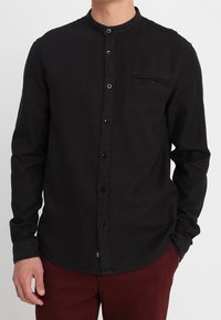 Svart långärmad skjorta med mandarin-krage, frontknäppning, en bröstficka och en texturerad tygkvalitet. Matchad med vinröda byxor.