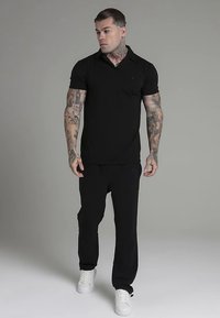 Musta lyhythihainen poolopaita, jossa on kaulus, yhdistetty mustiin housuihin ja valkoisiin lenkkareihin. Paidassa on rintatasku ja sileä kankaan pinta.