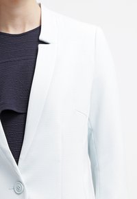 Detailní záběr na osobu v světle modré saku přes strukturovaný tmavě námořnický top, přičemž jsou vidět podrobnosti klopy a knoflíků saku.
