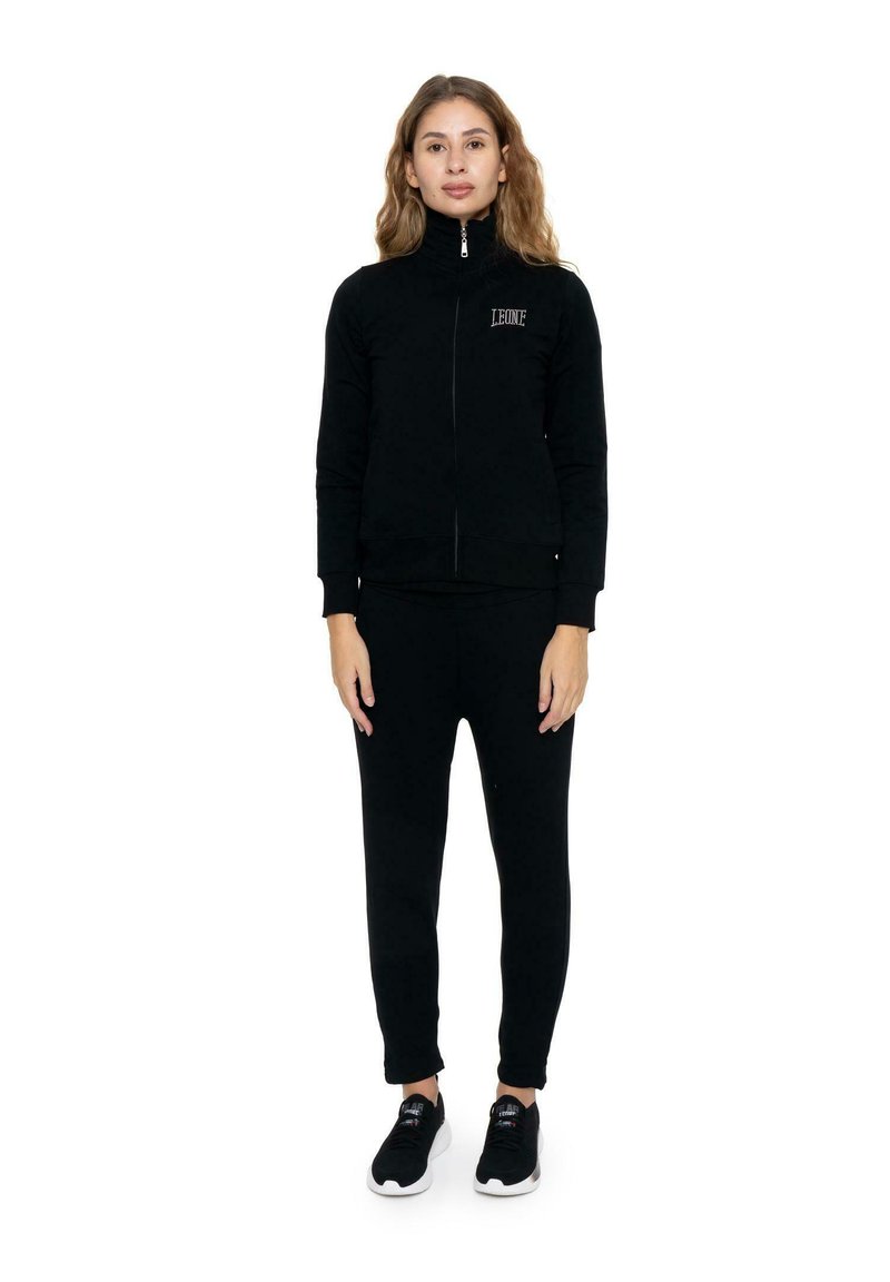 Une femme se tient devant un fond blanc, vêtue d’une veste noire zippée, d’un pantalon assorti et de baskets noires.