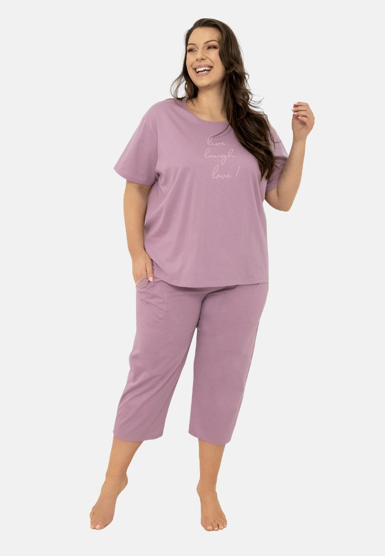 Uśmiechnięta kobieta w lawendowym dresie z napisem "żyj... śmiej się... kochaj!" na koszulce, stojąca boso na białym tle.