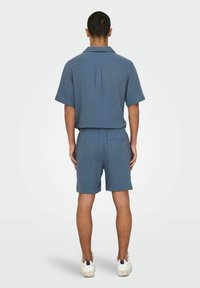 Blauwe shirt met korte mouwen en bijpassende korte broek van textuurstof. Beschikt over een rugplooi en een minimalistisch ontwerp; gecombineerd met witte sneakers.