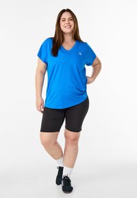 Blaues, athletisches T-Shirt mit V-Ausschnitt, kombiniert mit schwarzen Radlershorts. Das T-Shirt hat einen lockeren Schnitt, die Shorts sind eng anliegend. Schwarze Sportschuhe werden getragen.
