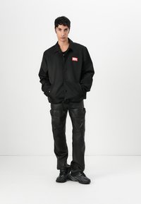 Svart, knappet jakke med en rød logo, kombinert med svarte lær cargo-bukser med lommer og snøring. Svarte sneakers fullfører stilen.