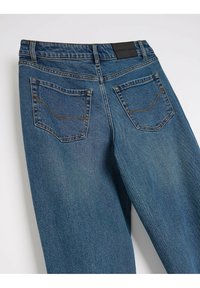 Jeans dżinsowe w średnim niebieskim odcieniu, z dwoma tylnymi kieszeniami z kontrastowym przeszyciem oraz czarną metką na pasku.
