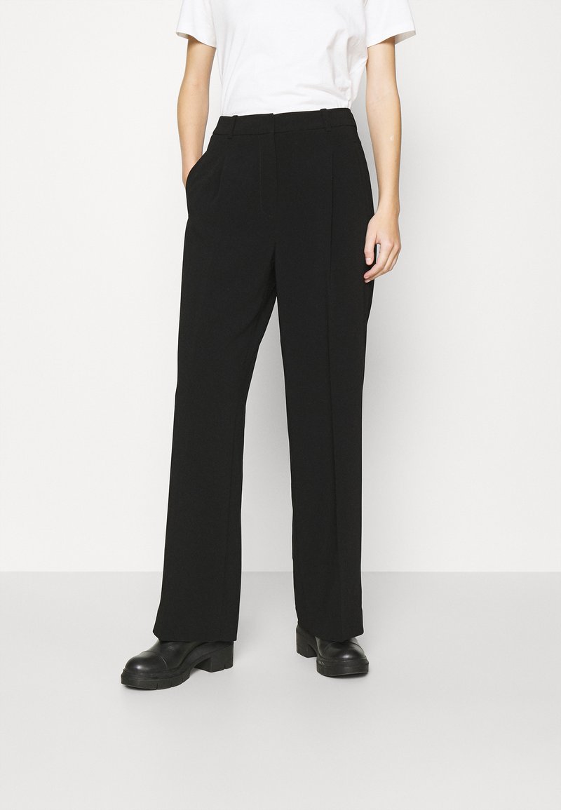 Zwarte, hoge taille broek met een rechtlopende pasvorm, voorzien van zijzakken en een gladde textuur. Gecombineerd met zwarte schoenen.