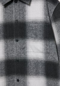 Cămașă de flanel în carouri gri și alb, cu guler cu nasturi, buzunar frontal și material texturat, având o combinație de pătrate întunecate și deschise.