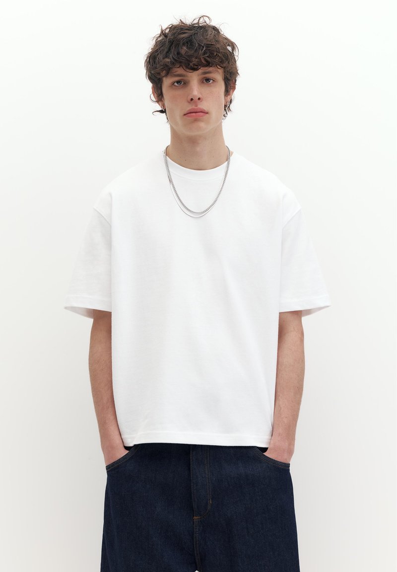 Junger Mann mit lockigem Haar, der ein schlichtes weißes, übergroßes T-Shirt, dunkle Jeans und eine silberne Kettenhalskette trägt, steht mit den Händen in den Taschen.