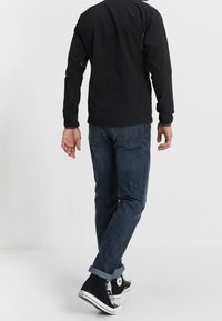 Svart långärmad skjorta, blå denimjeans, uppvikta ärmar och svarta höga sneakers, sedd bakifrån. Enfärgad bakgrund.