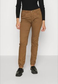 Pessoa a usar calças castanhas justas, uma blusa preta de manga longa e sapatos pretos sem atacadores, de pé contra um fundo claro e liso.