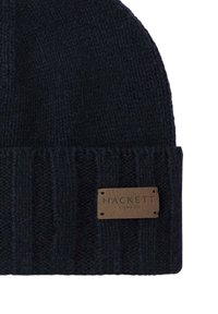 Knitted navy blue beanie with ribbed texture, featuring a brown leather tag embossed with "HACKETT LONDON" attached to the cuff.
