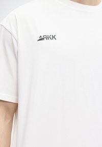 Λευκό κοντομάνικο μπλουζάκι με μαύρο λογότυπο "ARKK" στην πάνω αριστερή πλευρά του στήθους, φορεμένο από ένα άτομο που φαίνεται από τους ώμους μέχρι το μέσο του κορμού.