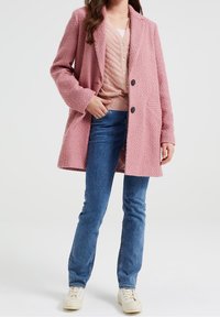 Rosa texturerad kappa med notchad krage, två svarta knappar och sidofickor, bärs över en ljusrosa lager-top och blå jeans.