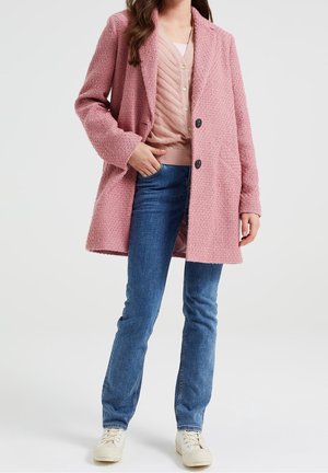 Abrigo texturizado rosa con cuello de muesca, dos botones negros y bolsillos laterales, usado sobre una blusa de capas rosa claro y jeans azules.