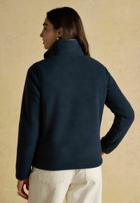 Žena oblečená v námorní modré fleecovej bunde s nápisom "JOULES" embosovaným na golieri, v kombinácii so svetlo béžovými nohavicami, stojaca pred béžovým pozadím.