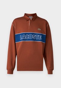 Dlhý rukáv polo tričko v hrdzavej hnedej farbe s modrým horizontálnym pruhom, na ktorom je biele písmo "LACOSTE". Obsahuje golier a detail zeleného loga.