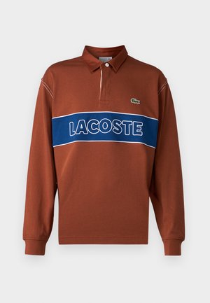 Polo a maniche lunghe di colore marrone ruggine con una striscia orizzontale blu e il testo bianco "LACOSTE". Include un colletto e un dettaglio del logo verde.
