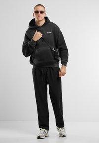 Crna oversized dukserica s kenguruh džepom, matching crne trenirke i tenisice s srebrnim detaljima. Uključuje sunčane naočale i malu torbu preko tijela.