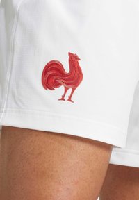 Shorts blancs arborant un logo de coq rouge brodé sur le côté gauche, mettant en avant un tissu texturé et un design épuré.