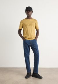 Mustard yellow t-shirt with a textured "BOSS" logo, paired with blue denim jeans. Model wears a black cap and black sneakers.