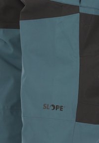 Vattentåliga tealfärgade och svarta skidbyxor med en slät yta och varumärket "SLOPE" tryckt i svart. Lapptäcktsdesign.