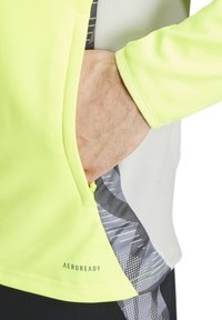 Leuchtend gelbes Langarmshirt mit grauem Einsatz, ausgestattet mit einer seitlichen Tasche mit Reißverschluss, texturierten Grafiken und "AEROREADY"-Markierung.