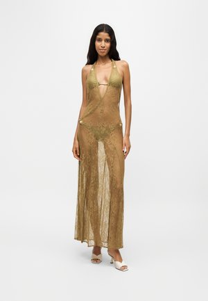 Femme portant une robe maxi en dentelle vert olive transparente sur un bikini assorti avec des talons blancs ouverts, se tenant devant un fond blanc uni.