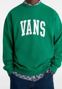 Felpa verde con logo bianco "VANS"; scollatura a girocollo, maniche lunghe, tessuto morbido, indossata sopra una camicia a scacchi grigia, abbinata a jeans blu.