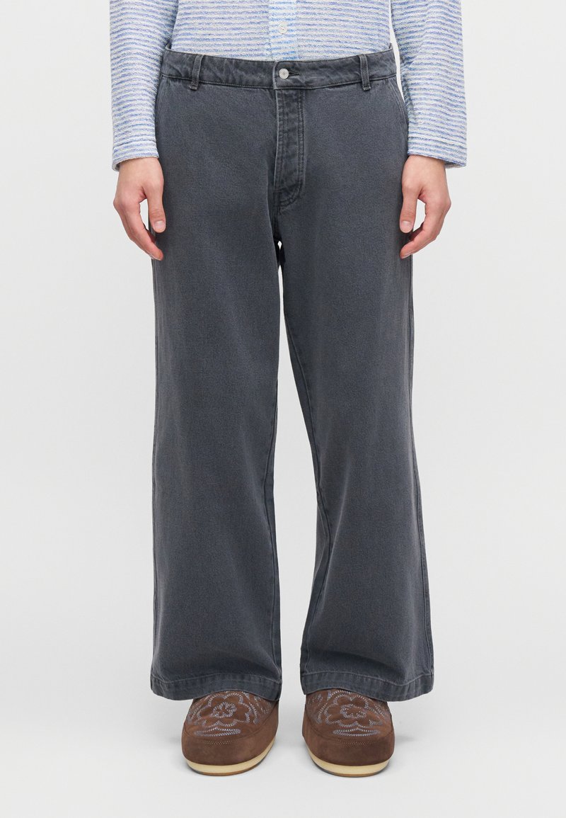 Τζιν παντελόνια σε στυλ wide-leg, σκούρου γκρι χρώματος με λεία υφή και θήκες για ζώνη. Συνδυάζονται με καφέ παπούτσια με σχέδιο, σε απλό φόντο.