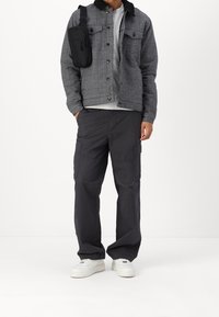 Man med grå rutiga jacka, ljusgrå skjorta, svarta cargobyxor, vita sneakers och svart axelremsväska mot vit bakgrund.