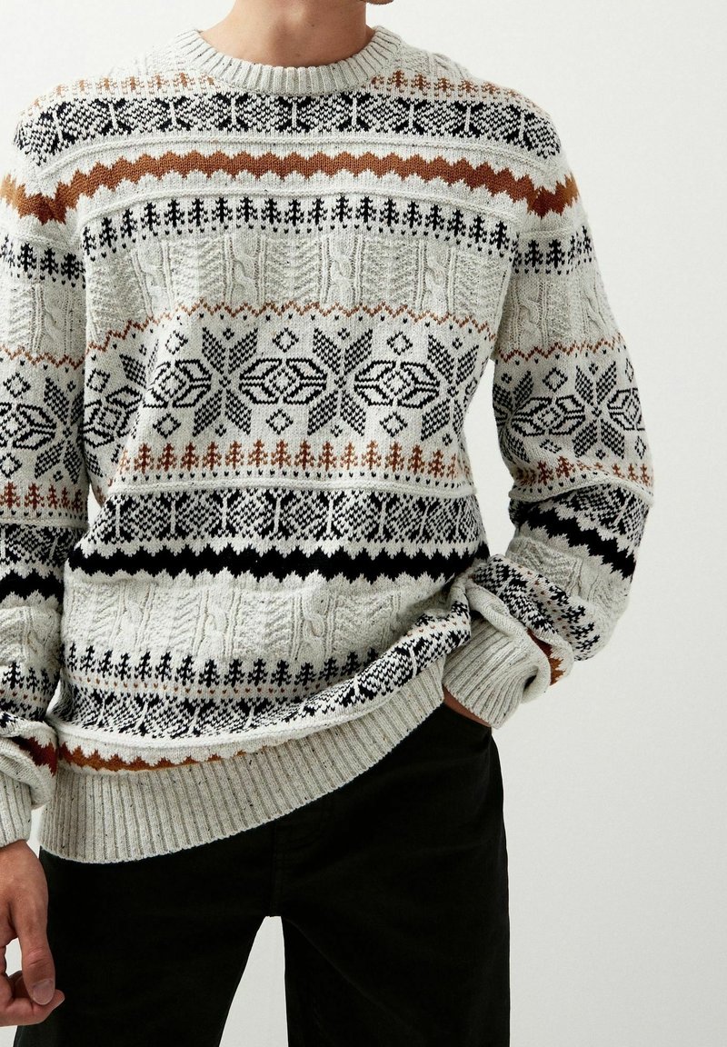 Homme portant un pull en maille gris avec des motifs nordiques noirs, marron et blancs et un pantalon noir, posant avec une main dans la poche.