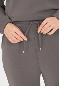 Graue Jogginghosen mit einem Kordelzugbund, goldenen Hardware-Akzenten und einem weichen, strukturierten Stoff. Fokus auf dem Bund und dem Kordelzugdetail.