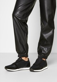 Black athletic sneakers with a mesh upper and suede accents, featuring a white speckled sole and cushioned ankle support. Paired with black joggers.