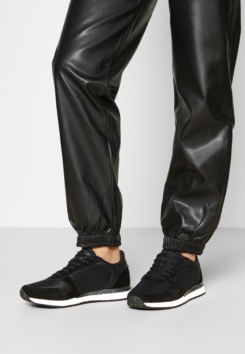 Black athletic sneakers with a mesh upper and suede accents, featuring a white speckled sole and cushioned ankle support. Paired with black joggers.