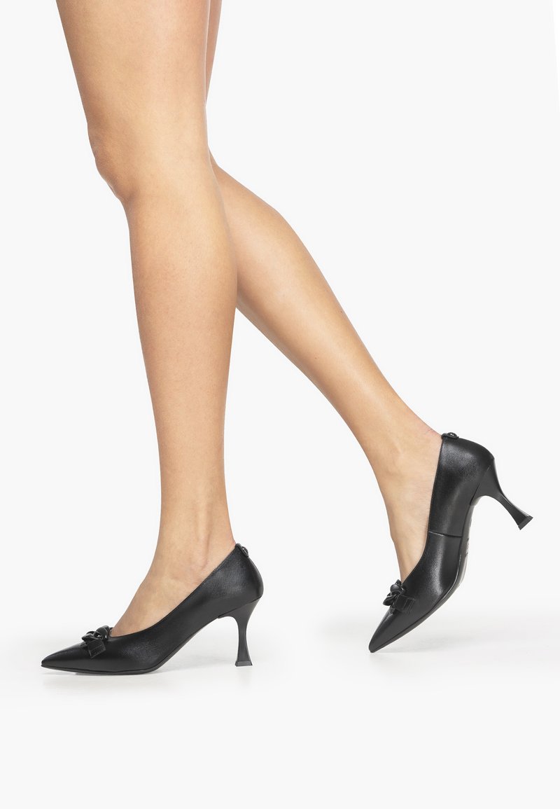 Black leather high-heeled shoes with a pointed toe and decorative bow detail on the front. Stiletto heel and smooth texture.