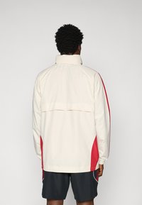 Cream-colored jacket with red side accents, tall collar, and elastic cuffs. Lightweight design; back features a horizontal panel.