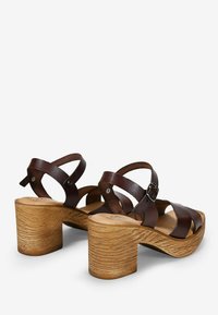Sandalias de cuero marrón con correas y tacón de madera gruesa. Presentan correas ajustables y una plantilla con curvatura para mayor soporte.