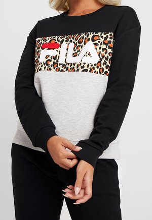 Vrouw draagt een zwart-grijze FILA-trui met luipaardprintdetail over de borst, terwijl ze de mouwmanchet van haar linkerarm aanpast.