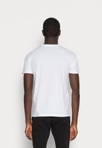 Bílý bavlněný tričko s kulatým výstřihem a krátkými rukávy, zobrazené zezadu, v kombinaci s černými kalhotami. Hladká textura.