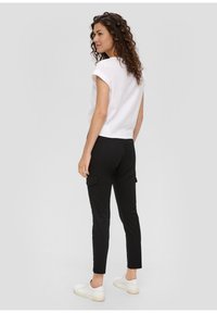 Witte korte mouwen top, zwarte getailleerde cargo broek met zijzakken en witte sneakers, gezien vanuit de rug tegen een neutrale achtergrond.
