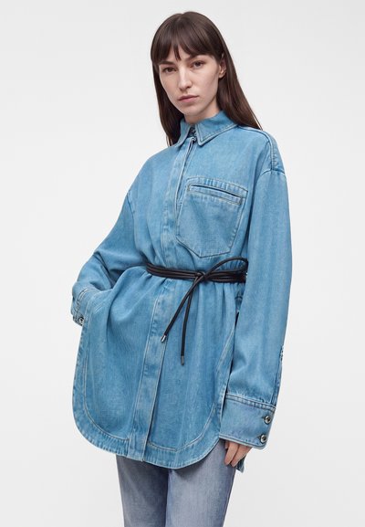Jeune femme aux cheveux bruns et lisses portant une veste en jean bleue surdimensionnée ceinturée d'un cordon noir fin et un jean bleu clair sur fond blanc.