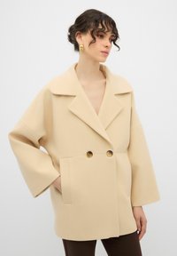 Cappotto beige oversize con ampio risvolto, due grandi bottoni e tasche laterali. Tessuto morbido con maniche larghe, progettato per una vestibilità rilassata.