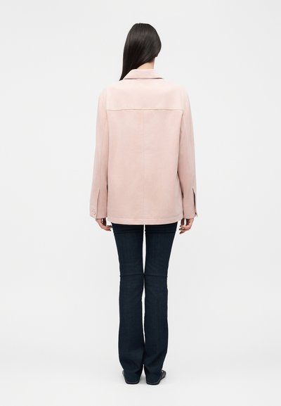 Γυναίκα με μακριά σκούρα μαλλιά, που φοράει ένα ανοιχτό ροζ μπουφάν και σκούρο μπλε τζιν, στέκεται με την πλάτη προς ένα απλό λευκό φόντο.