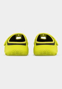 Φωτεινές κίτρινες σανδάλες χωρίς λουράκια με μαύρη εσωτερική επένδυση, απεικονιζόμενες από πίσω, σε απλό ανοιχτό γκρι φόντο.