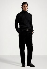 Μαύρο πλεκτό πουλόβερ ζιβάγκο με σχέδιο πλεξίδας, συνδυασμένο με μαύρο βελούδινο παντελόνι και μαύρα παπούτσια, τονισμένο με λεπτή μαύρη ζώνη.