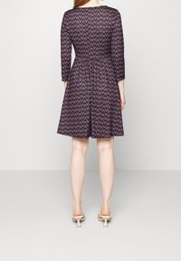 Czarna sukienka w fioletowy kwiatowy wzór, z rękawami 3/4, wcięciem w talii, rozkloszowanym dołem, noszona z beżowymi sandałami na obcasie.