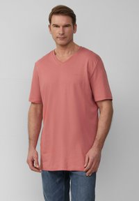 Jasnoróżowa koszulka typu v-neck z krótkim rękawem, wykonana z bawełny. Posiada subtelne logo na klatce piersiowej oraz luźny krój, w zestawie z niebieskimi dżinsami.