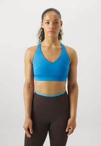 Αθλητικό σουτιέν σε φωτεινό μπλε, με σχέδιο V-λαιμόκοψης και φαρδιά τιράντες. Συνδυασμένο με καφέ κολάν ψηλής μέσης με μπλε λάστιχο.