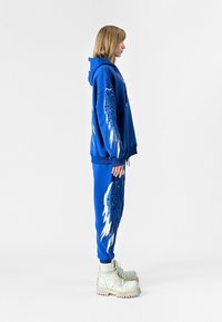 Sudadera azul y pantalones de chándal con diseños de alas en blanco y azul. El conjunto se complementa con botas en color marfil con suelas rugosas.